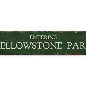 以下が含まれることがあります： 「ENTERING YELLOWSTONE PARK」の白い文字が書かれた、長方形の、使い古された緑色の金属看板。看板は、錆びた縁と目に見える摩耗があり、風化した外観をしています。