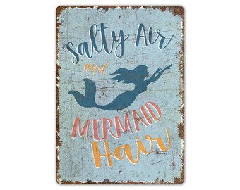 May include: A blue and white metal sign with a distressed look. The sign says "Salty Air and Mermaid Hair" with a mermaid silhouette in the middle.