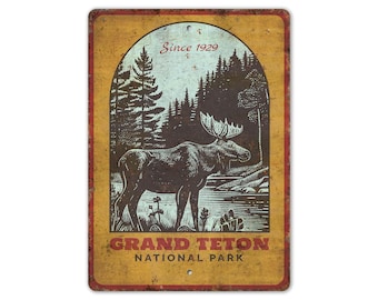 May include: Aged metal sign depicting a moose in a forest, with the words "Grand Teton National Park" and "Since 1929". The sign has a distressed look with a yellow and brown colour scheme, suitable for a rustic aesthetic.