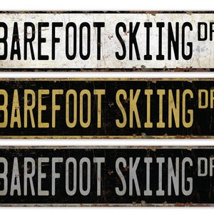 May include: Three vintage-style metal signs with the text "BAREFOOT SKIING DR" in black, gold, and gray lettering on a black, white, and black background.