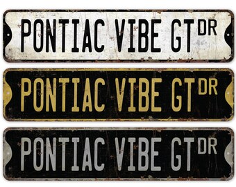 May include: Three vintage-style metal signs with the text "PONTIAC VIBE GT DR" in black, gold, and silver lettering on a black background.