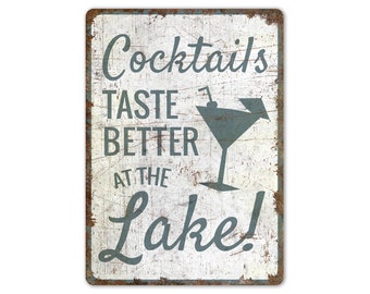 May include: A rectangular metal sign with a distressed white background and a rusty brown border. The sign features the text "Cocktails TASTE BETTER AT THE LAKE!" in a teal script font, along with a teal martini glass illustration.