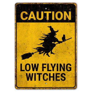 Puede incluir: Un letrero metálico rectangular con fondo amarillo y texto y gráficos negros. El letrero dice "CAUTION LOW FLYING WITCHES". Una silueta negra de una bruja en una escoba con un gato está en el centro. El letrero tiene un aspecto vintage.