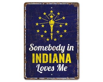 Może przedstawiać: Metalowy znak w kolorze niebieskim i żółtym z tekstem "Somebody in INDIANA Loves Me". Znak przedstawia flagę stanu Indiana z pochodnią i gwiazdami.
