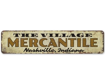 Op de afbeelding: Rechthoekig metalen bord met een verweerde, vintage uitstraling. Het bord toont de woorden "THE VILLAGE MERCANTILE" in bruine letters, met "Nashville, Indiana" eronder. De achtergrond is gebroken wit.