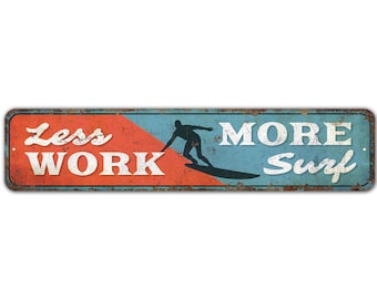 May include: A vintage-style metal sign with a blue and red background. The sign reads "Less Work More Surf" with a silhouette of a surfer on a wave.