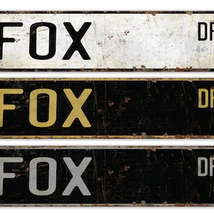 May include: Three vintage-style metal street signs with the text "FOX DR" on each. The signs are in different color combinations: white with black text, black with gold text, and black with silver text.