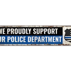 May include: A rusty metal sign with the text "WE PROUDLY SUPPORT OUR POLICE DEPARTMENT" in white letters on a black and blue background. A thin blue line American flag is on the right side of the sign.
