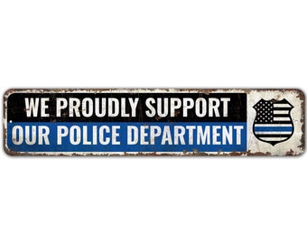 May include: A rusty metal sign with the text "WE PROUDLY SUPPORT OUR POLICE DEPARTMENT" in white letters on a black and blue background. A thin blue line American flag is on the right side of the sign.