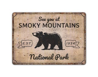 May include: Rectangular metal sign with a distressed finish, featuring the text "See you at SMOKY MOUNTAINS National Park." A black bear silhouette is centered above the words. The sign includes "EST" and "1934" in banner shapes.