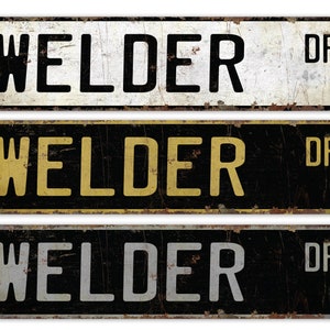 May include: Three vintage-style metal street signs with the text "Welder Dr." in black, gold, and silver lettering on a white, black, and black background, respectively.