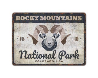 May include: Distressed metal sign with the text "Rocky Mountains" and "National Park Colorado, USA." The sign features a geometric ram's head. The sign includes the text "ES TD" and the year "1915". The sign has a rectangular shape.