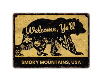 May include: A rectangular metal sign with a distressed finish. The sign features a black bear silhouette filled with floral designs. The words "Welcome, Y'all" are arched above the bear, and "SMOKY MOUNTAINS, USA" is below.