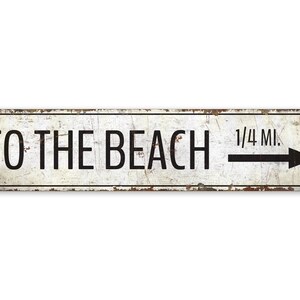 Könnte beinhalten: Weißes Metallschild mit schwarzem Text und einem nach rechts zeigenden Pfeil. Das Schild lautet "To the Beach 1/4 Mi."