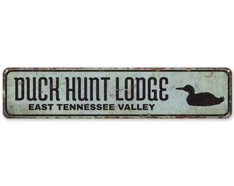 Może przedstawiać: Prostokątny metalowy znak o postarzanym, vintage wyglądzie. Znak zawiera słowa "DUCK HUNT LODGE" nad "EAST TENNESSEE VALLEY" w czarnych literach. Sylwetka kaczki znajduje się po prawej stronie. Tło jest jasnoniebieskie.