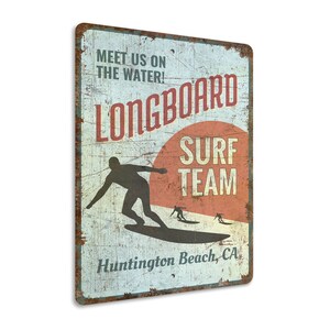 May include: Vintage-style metal sign with a distressed look. It features a silhouette of a surfer riding a longboard with the text "Longboard Surf Team" and "Huntington Beach, CA".