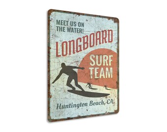 May include: Vintage-style metal sign with a distressed look. It features a silhouette of a surfer riding a longboard with the text "Longboard Surf Team" and "Huntington Beach, CA".