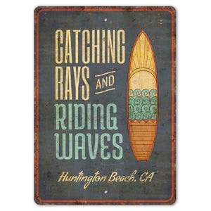 May include: A vintage-style metal sign with a blue background and a surfboard graphic. The text on the sign reads "Catching Rays and Riding Waves Huntington Beach, CA".