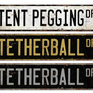 Può includere: Tre cartelli stradali in metallo in stile vintage con angoli arrotondati e bordi leggermente arrugginiti. Il cartello superiore è bianco con testo nero che recita "TENT PEGGING DR". Il cartello centrale è nero con testo dorato che recita "TETHERBALL DR". Il cartello inferiore è nero con testo argentato che recita "TETHERBALL DR".
