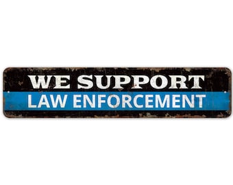 May include: A black and blue metal sign with the text "WE SUPPORT LAW ENFORCEMENT" in white letters. The sign has a distressed, rusty look.