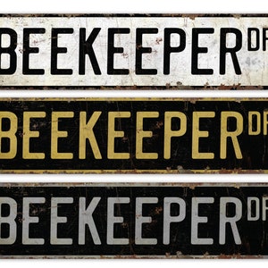 Könnte beinhalten: Drei Schilder im Vintage-Stil aus Metall mit dem Text "BEEKEEPER DR" in schwarzen Buchstaben auf weißem, gelbem und grauem Hintergrund. Die Schilder haben ein abgenutztes, rostiges Aussehen.
