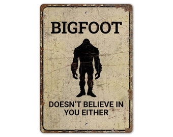 Op de afbeelding: Een vintage metalen bord met een verweerde achtergrond. Het bord heeft een zwart silhouet van Bigfoot en de tekst "BIGFOOT GELOOFT NIET IN JOU."