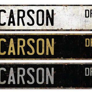 Puede incluir: Tres señales de calle de metal de estilo vintage con el texto "CARSON DR" en letras negras, doradas y plateadas sobre un fondo blanco y negro.