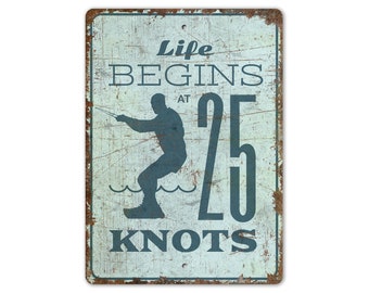 May include: A vintage-style metal sign with a distressed white background and blue text. The sign features a silhouette of a person waterskiing and the text "Life Begins at 25 Knots".
