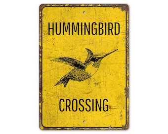 Puede incluir: Un letrero de metal amarillo con un acabado desgastado. El letrero dice "Hummingbird Crossing" con una silueta negra de un colibrí en vuelo.