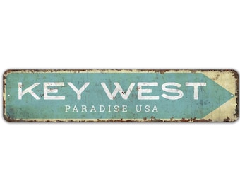 May include: A vintage-style metal sign with a blue background and white text that reads "KEY WEST PARADISE USA". The sign has a distressed, rusty look and a white arrow pointing to the right.