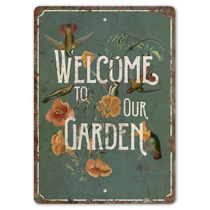 Puede incluir: Un letrero de metal de estilo vintage con fondo azul y bordes oxidados. El letrero presenta un mensaje de bienvenida con el texto "Bienvenido a nuestro jardín" e ilustraciones de colibríes y flores.