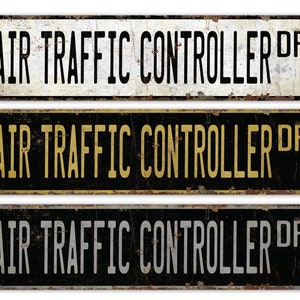 May include: Three vintage-style metal signs with the text "AIR TRAFFIC CONTROLLER DR". The signs are rectangular with rounded corners and a distressed, rusty finish. The top sign is white with black text, the middle is black with yellow text, and the bottom is black with white text.