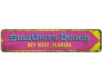 Può includere: Un cartello in metallo in stile vintage con bordo giallo e sfondo rosa. Il cartello recita "Smathers Beach KEY WEST, FLORIDA" con una freccia blu che punta a destra.