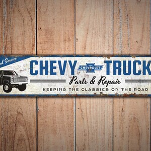 K&ouml;nnte beinhalten: Ein rechteckiges Metallschild mit abgenutztem Finish, das die Worte "CHEVY TRUCKS" in Blau und eine schwarze LKW-Illustration zeigt. Das Schild enth&auml;lt auch den Text "Parts & Repair" und "KEEPING THE CLASSICS ON THE ROAD."