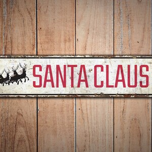 Op de afbeelding: Een wit metalen bord met rode tekst die "SANTA CLAUS LN" zegt. Het bord heeft een verweerde uitstraling en toont een silhouet van de kerstman in zijn slee met rendieren.