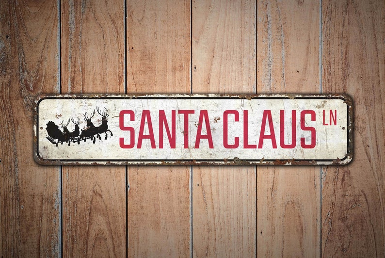 Op de afbeelding: Een wit metalen bord met rode tekst die "SANTA CLAUS LN" zegt. Het bord heeft een verweerde uitstraling en toont een silhouet van de kerstman in zijn slee met rendieren.