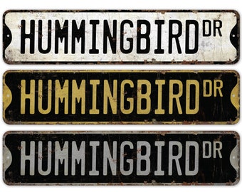 Puede incluir: Tres letreros de metal de estilo vintage con el texto "HUMMINGBIRD DR" en letras negras, amarillas y blancas sobre fondo negro. Los letreros tienen un aspecto desgastado y oxidado.
