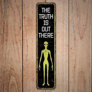 Puede incluir: Un letrero rectangular de metal con el texto "THE TRUTH IS OUT THERE" en letras blancas. Debajo, una figura de alien&iacute;gena verde. El letrero tiene un fondo negro con un aspecto vintage y desgastado. Montado sobre una superficie de madera.
