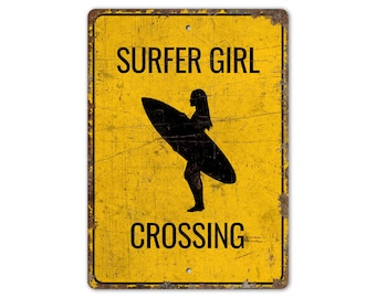 May include: Yellow metal sign with a distressed look. The sign reads "Surfer Girl Crossing" with a silhouette of a surfer girl holding a surfboard.