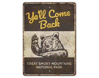 May include: A vintage-style metal sign with a brown background and white lettering. The sign says "Ya'll Come Back" in yellow letters and features a black and white illustration of a bear waving. The sign also says "Great Smoky Mountains National Park" in white letters.