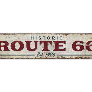 以下が含まれることがあります： 白地に赤い文字で「Historic Route 66 Est. 1926」と書かれた、ビンテージスタイルの金属製の看板。看板は、使い古された、さびたような外観をしています。