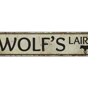 May include: Rectangular metal sign with the words "WOLF'S LAIR" in black, distressed font. A black silhouette of a wolf is on the right. The sign has a weathered, vintage appearance with rust-colored edges.