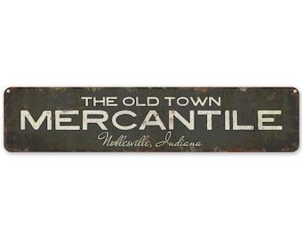 Op de afbeelding: Een vintage metalen bord met een rustieke groene achtergrond en witte letters. Het bord heeft de tekst "The Old Town Mercantile" met de tekst "Noblesville, Indiana" in een kleiner lettertype eronder.