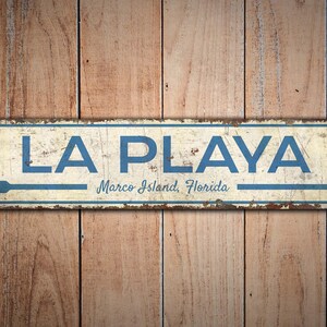 Puede incluir: Un letrero met&aacute;lico rectangular con las palabras "LA PLAYA" en letras azules, con "Marco Island, Florida" debajo. El letrero tiene una flecha que apunta hacia la derecha y est&aacute; montado sobre un fondo de madera.