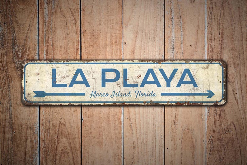 Puede incluir: Un letrero met&aacute;lico rectangular con las palabras "LA PLAYA" en letras azules, con "Marco Island, Florida" debajo. El letrero tiene una flecha que apunta hacia la derecha y est&aacute; montado sobre un fondo de madera.