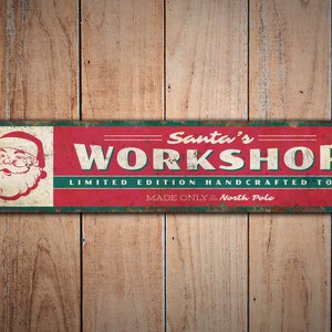 May include: Red and green metal sign with the text "Santa's Workshop" and "Limited Edition Handcrafted Toys Made Only at the North Pole". A cartoon image of Santa Claus is on the left side of the sign.