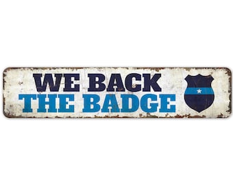 May include: A white metal sign with blue and black text that reads "We Back The Badge". A blue and black police badge with a white star is on the right side of the sign.