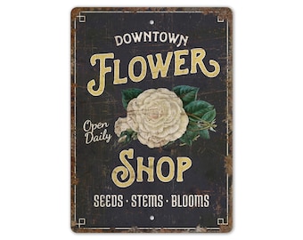 May include: A distressed metal sign advertising a flower shop. The sign reads "Downtown Flower Shop" in gold lettering, with a white rose illustration. It also includes "Open Daily" and "Seeds, Stems, Blooms".