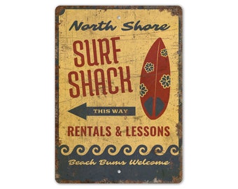 May include: A vintage-style metal sign with a yellow background and blue and red accents. The sign reads "North Shore Surf Shack" with an arrow pointing to the right and the text "This Way". Below the arrow is the text "Rentals & Lessons" and at the bottom of the sign is the text "Beach Bums Welcome".