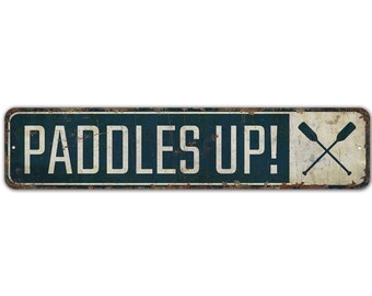 以下が含まれることがあります： 「PADDLES UP!」という文字と、角に交差したパドルが描かれた青と白の金属製の看板です。